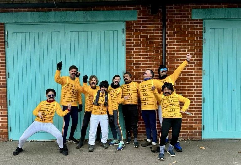 Mixed masters eight crew posing in front of Caius boat house doors dressed for their Bohemian Rhapsody theme in yellow tops and fake moustaches