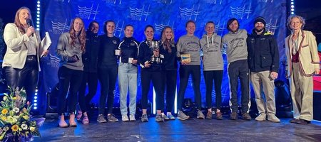 The winning Cantabs crew posing on a stage with their medals, trophy and plaque. They are joined by Camilla Hadland who was MC for the event, and Annamarie Phelps who presented them with their prizes.