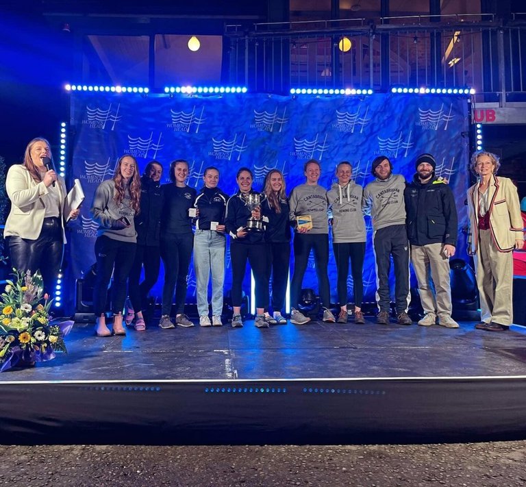 The winning Cantabs crew posing on a stage with their medals, trophy and plaque. They are joined by Camilla Hadland who was MC for the event, and Annamarie Phelps who presented them with their prizes.